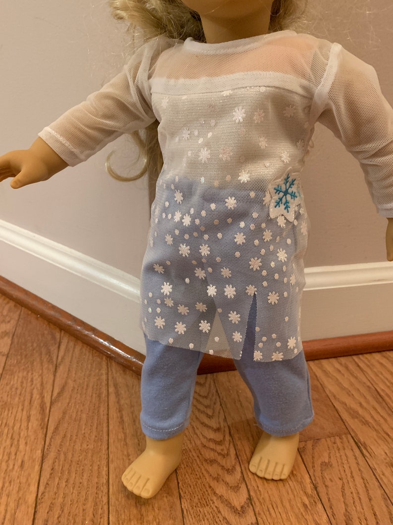 May include: A doll wearing a two-piece outfit. The top is a white mesh long-sleeve shirt with a snowflake pattern over a blue base. The outfit includes blue pants. A blue and white snowflake patch is on the shirt.