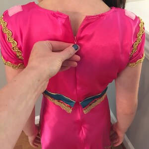 May include: A pink dress with gold trim and a zipper in the back. The dress has a blue and gold sash around the waist.