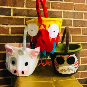 Puede incluir: Tres cubos de truco o trato de Halloween de fieltro con forma de gallo, cerdo y un personaje de Moana. El cubo de gallo es amarillo, rojo y azul. El cubo de cerdo es blanco con manchas grises. El cubo del personaje de Moana es marr&oacute;n con detalles negros y blancos.