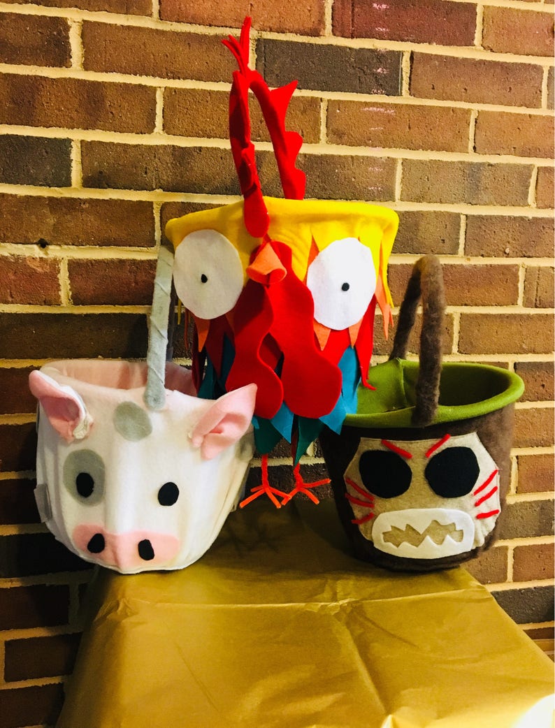 Puede incluir: Tres cubos de truco o trato de Halloween de fieltro con forma de gallo, cerdo y un personaje de Moana. El cubo de gallo es amarillo, rojo y azul. El cubo de cerdo es blanco con manchas grises. El cubo del personaje de Moana es marr&oacute;n con detalles negros y blancos.