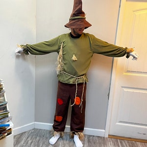 K&ouml;nnte beinhalten: Eine Person, die sich als Vogelscheuche f&uuml;r Halloween verkleidet hat. Sie tr&auml;gt einen braunen, spitzen Hut, ein gr&uuml;nes Langarmshirt, eine braune Hose mit orangefarbenen und roten Flecken und wei&szlig;e Socken.