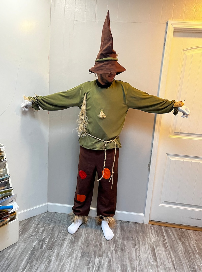 K&ouml;nnte beinhalten: Eine Person, die sich als Vogelscheuche f&uuml;r Halloween verkleidet hat. Sie tr&auml;gt einen braunen, spitzen Hut, ein gr&uuml;nes Langarmshirt, eine braune Hose mit orangefarbenen und roten Flecken und wei&szlig;e Socken.