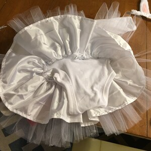 May include: White tulle tutu skirt with a white satin overlay and a white bodysuit. The skirt is gathered at the waist and has a ruffled edge. The bodysuit has a scoop neck and sleeveless design.