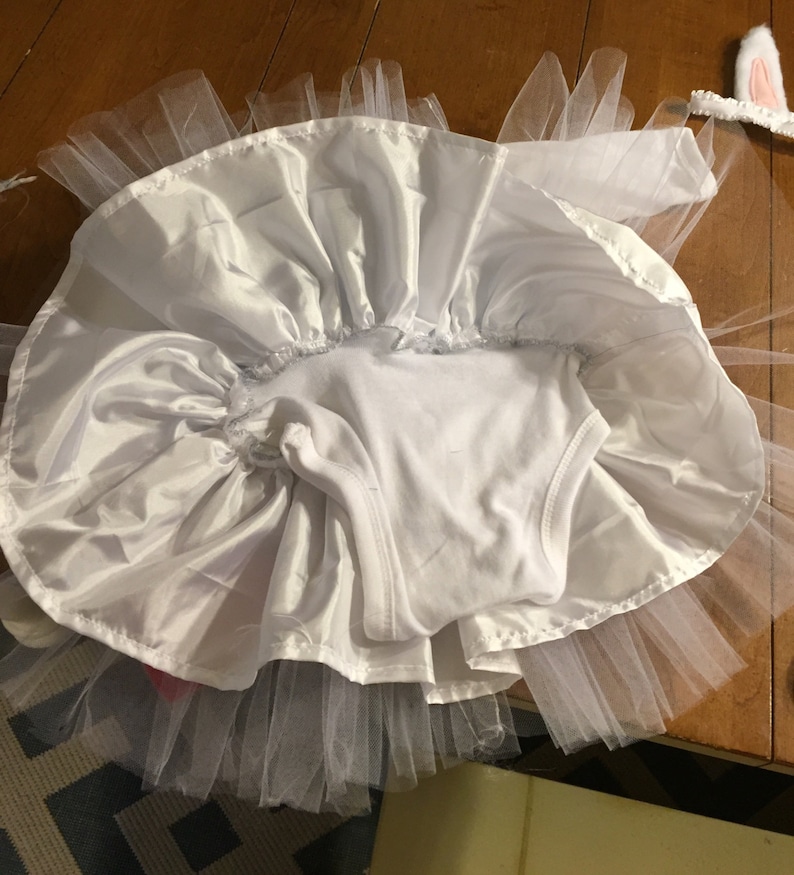 May include: White tulle tutu skirt with a white satin overlay and a white bodysuit. The skirt is gathered at the waist and has a ruffled edge. The bodysuit has a scoop neck and sleeveless design.