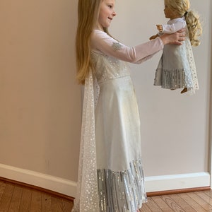 Puede incluir: Una ni&ntilde;a peque&ntilde;a con un vestido blanco y plateado con una capa larga y transparente sostiene una mu&ntilde;eca con un vestido similar. El vestido tiene un ribete plateado brillante y la capa est&aacute; salpicada de copos de nieve blancos.