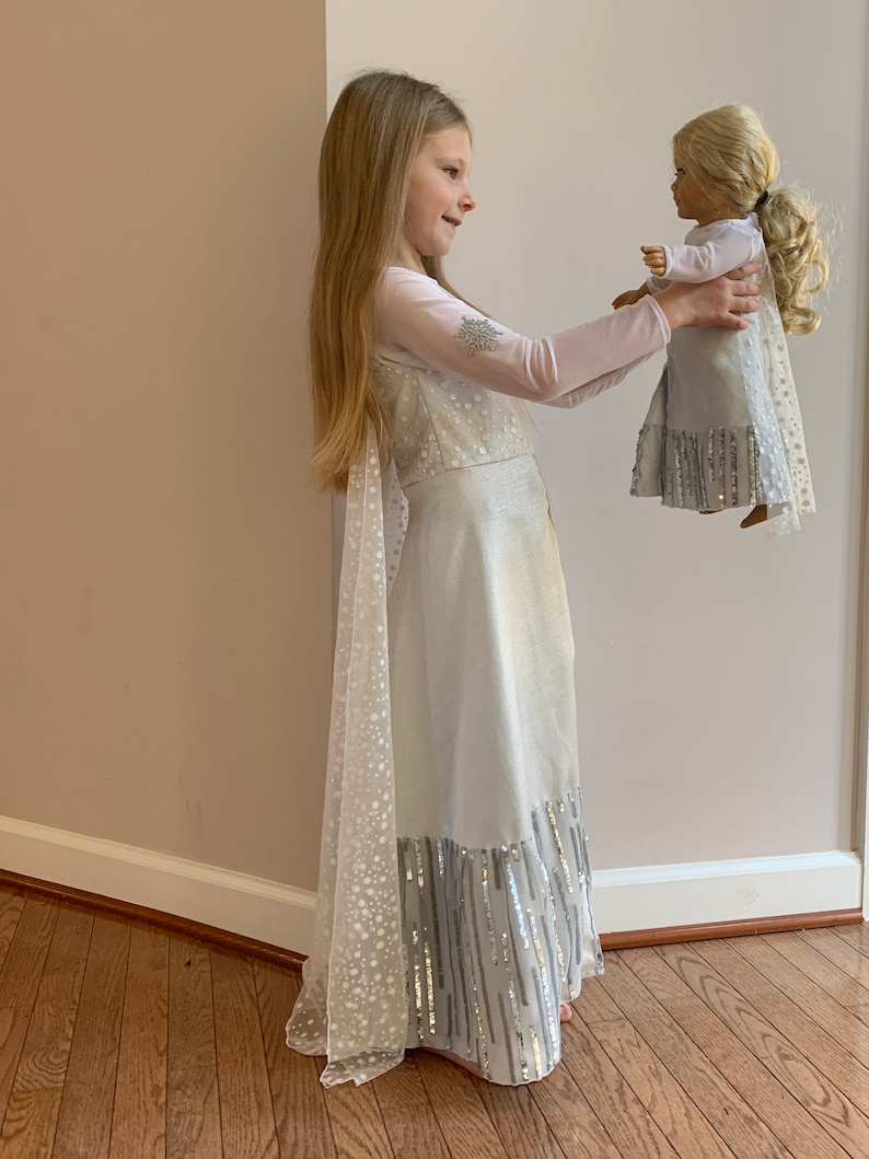 Puede incluir: Una ni&ntilde;a peque&ntilde;a con un vestido blanco y plateado con una capa larga y transparente sostiene una mu&ntilde;eca con un vestido similar. El vestido tiene un ribete plateado brillante y la capa est&aacute; salpicada de copos de nieve blancos.
