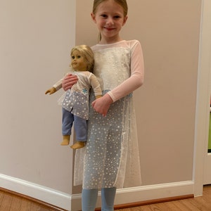 May include: A young girl wearing a white and light blue dress with a snowflake pattern, holding a doll dressed in a matching outfit. The doll has blonde hair and is wearing a light blue outfit with a snowflake design. The girl is also wearing light blue leggings.
