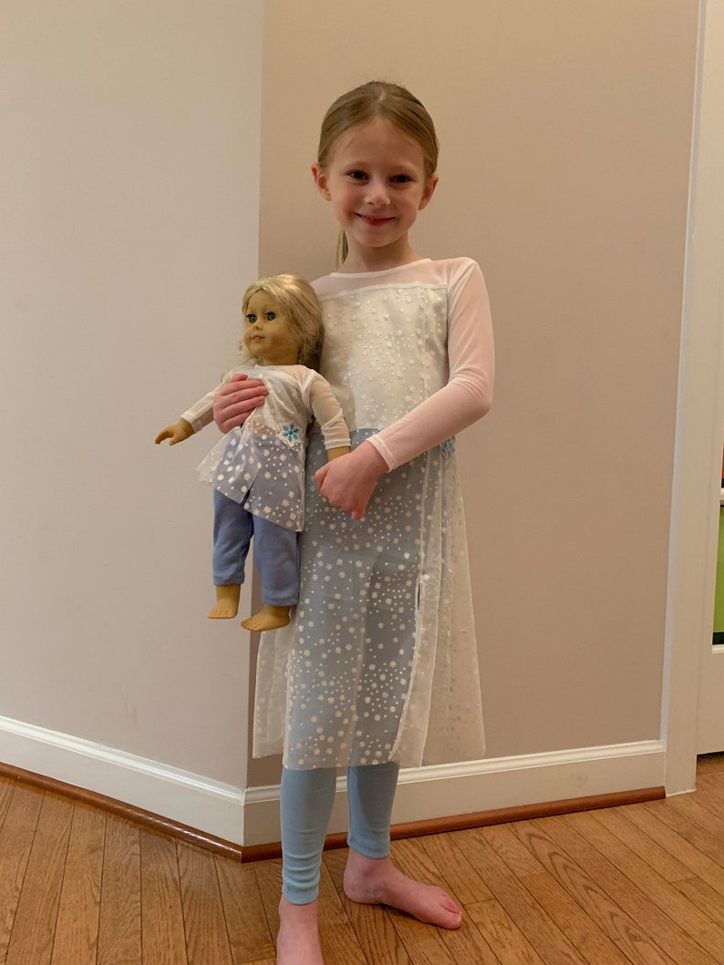 May include: A young girl wearing a white and light blue dress with a snowflake pattern, holding a doll dressed in a matching outfit. The doll has blonde hair and is wearing a light blue outfit with a snowflake design. The girl is also wearing light blue leggings.