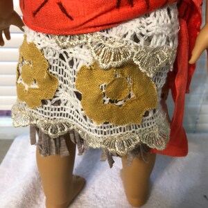 May include: A doll wearing a red and white outfit with a brown and gold skirt. The skirt has a crocheted design and a fringe.