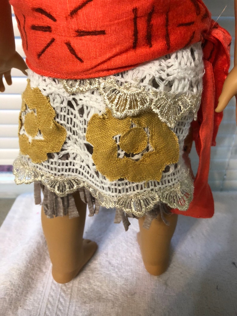 May include: A doll wearing a red and white outfit with a brown and gold skirt. The skirt has a crocheted design and a fringe.