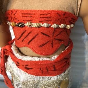 May include: A red and white doll outfit with a brown top and a white skirt with a crocheted lace trim. The outfit has a red sash with a brown pattern and a shell necklace.