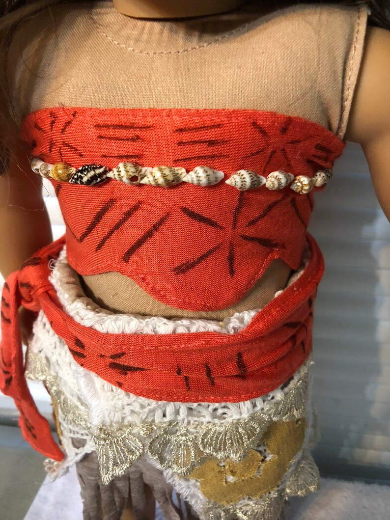 May include: A red and white doll outfit with a brown top and a white skirt with a crocheted lace trim. The outfit has a red sash with a brown pattern and a shell necklace.