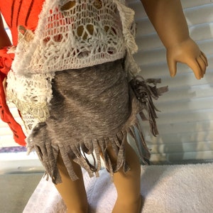 May include: A doll wearing a coral and white crochet top with a brown fringed skirt.