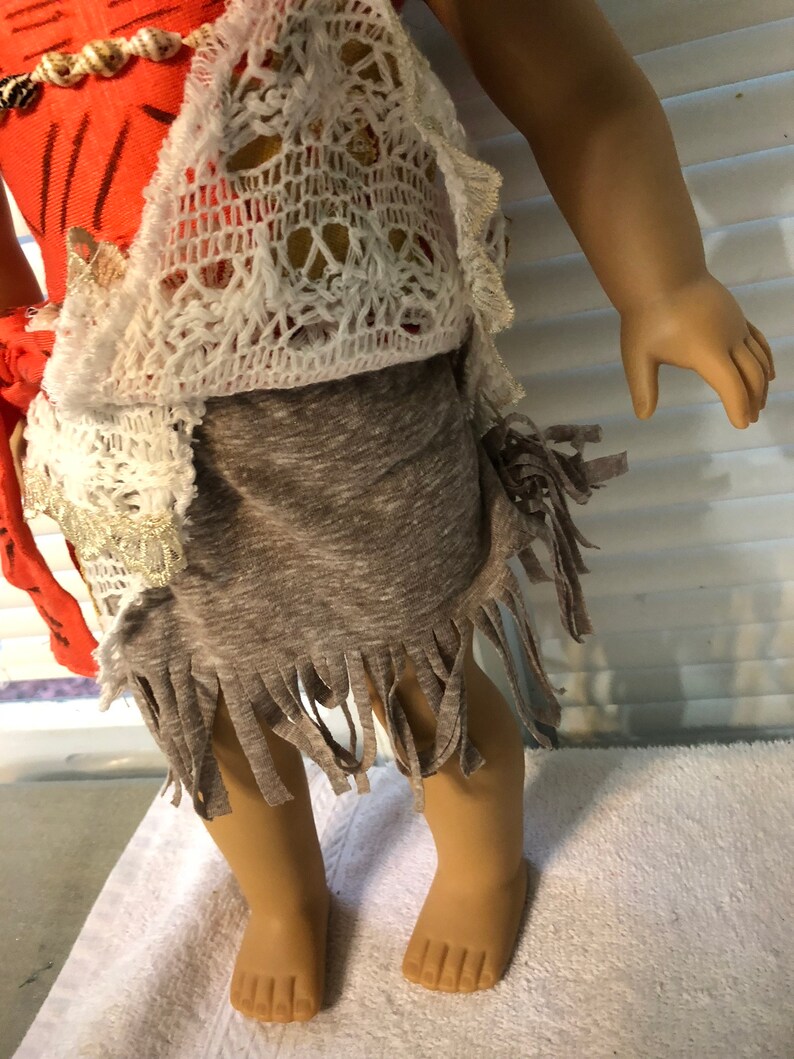 May include: A doll wearing a coral and white crochet top with a brown fringed skirt.