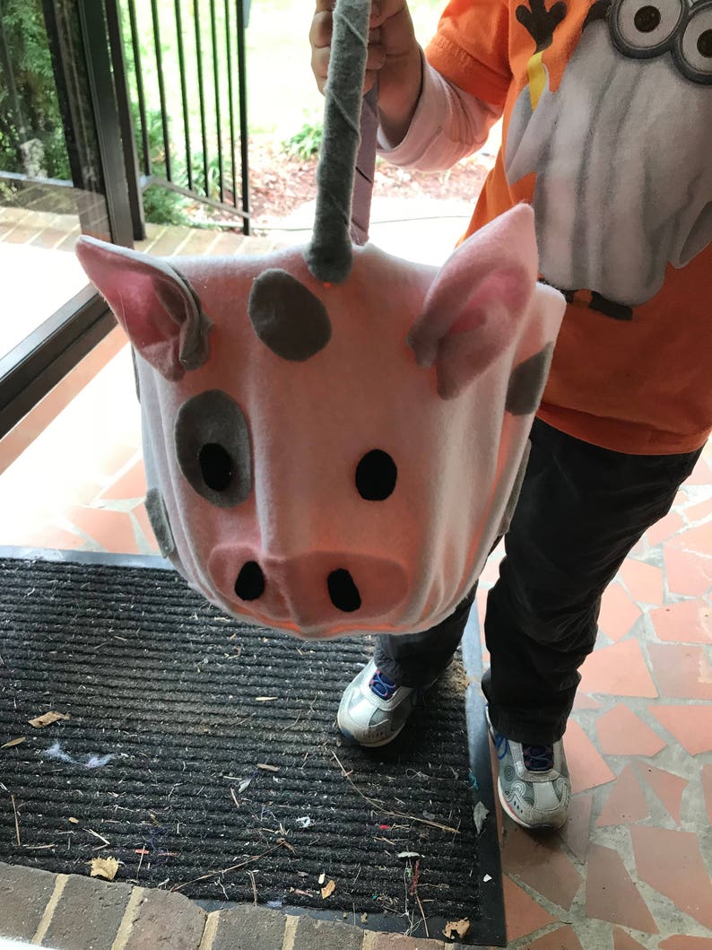 Puede incluir: Una bolsa de truco o trato en forma de cerdo rosa con manchas grises y ojos negros. La bolsa tiene un asa a rayas grises y blancas.