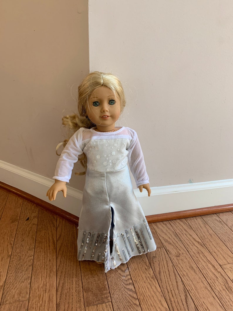 Puede incluir: Una mu&ntilde;eca con un vestido blanco de manga larga con una falda plateada y lentejuelas. La mu&ntilde;eca tiene el pelo rubio y los ojos azules.