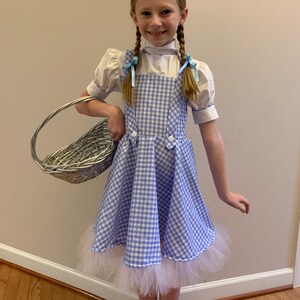 K&ouml;nnte beinhalten: Ein junges M&auml;dchen, das als Dorothy aus dem Zauberer von Oz verkleidet ist. Sie tr&auml;gt ein blaues und wei&szlig;es Gingham-Kleid mit einem wei&szlig;en Petticoat, eine wei&szlig;e Bluse mit Puff&auml;rmeln und blaue Schleifen im Haar. Sie h&auml;lt einen Weidenkorb.