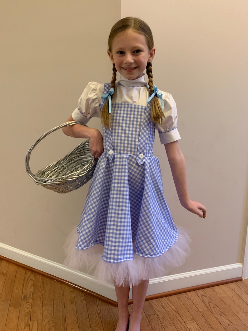 K&ouml;nnte beinhalten: Ein junges M&auml;dchen, das als Dorothy aus dem Zauberer von Oz verkleidet ist. Sie tr&auml;gt ein blaues und wei&szlig;es Gingham-Kleid mit einem wei&szlig;en Petticoat, eine wei&szlig;e Bluse mit Puff&auml;rmeln und blaue Schleifen im Haar. Sie h&auml;lt einen Weidenkorb.