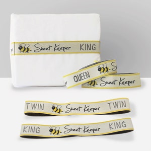 May include: Four beige and yellow elastic bands with a bee graphic and the text "Sheet Keeper" printed on them. The bands are labeled with the sizes "King", "Queen", "Twin", and "King".