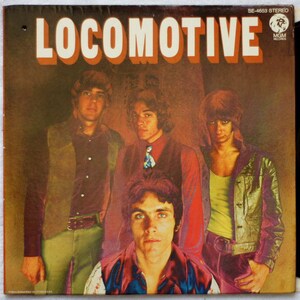 Op de afbeelding: Een vintage vinyl platenhoes met een band genaamd "Locomotive". De hoes toont vier mannen in een band, met een bruin, oranje en geel kleurenschema. De bandleden dragen casual kleding, waaronder een bruin vest, een paars shirt, een rood shirt en een blauw denim jack.