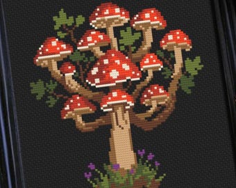 Op de afbeelding: Een ingelijst kruissteek borduurwerk met een gepixelde boom met een lichtbruine stam en takken, bekroond met meerdere rood-witte stippenpaddenstoelen, tegen een effen zwarte achtergrond in een eenvoudige zwarte lijst.