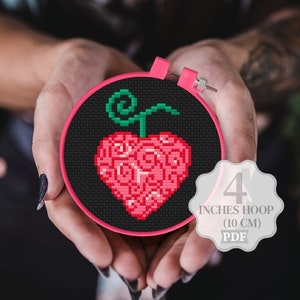 May include: A red and green heart-shaped fruit with swirls, cross-stitched on black fabric in a 4-inch embroidery hoop.  The hoop is pink and held by hands.