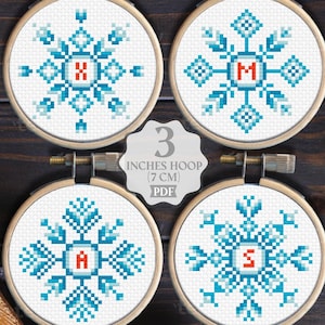 May include: Four circular wooden embroidery hoops containing cross-stitch snowflake patterns in shades of blue and white, each featuring a red letter in the center. Text reads: COMPATIBLE WITH PATTERN KEEPER, 3 INCHES HOOP (7 CM) PDF, WHOLE ALPHABET INCLUDED.