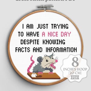 Op de afbeelding: PDF kruissteekpatroon met een humoristische quote: "I AM JUST TRYING TO HAVE A NICE DAY DESPITE KNOWING FACTS AND INFORMATION." Het ontwerp bevat een grijze opossum met roze accenten. De borduurring is 20 cm.