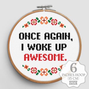 Op de afbeelding: Een borduurring met de tekst "ONCE AGAIN, I WOKE UP AWESOME." in zwart en rood. Het ontwerp bevat bloempatronen in oranje, rood en groen. De ring heeft een diameter van 15 cm.