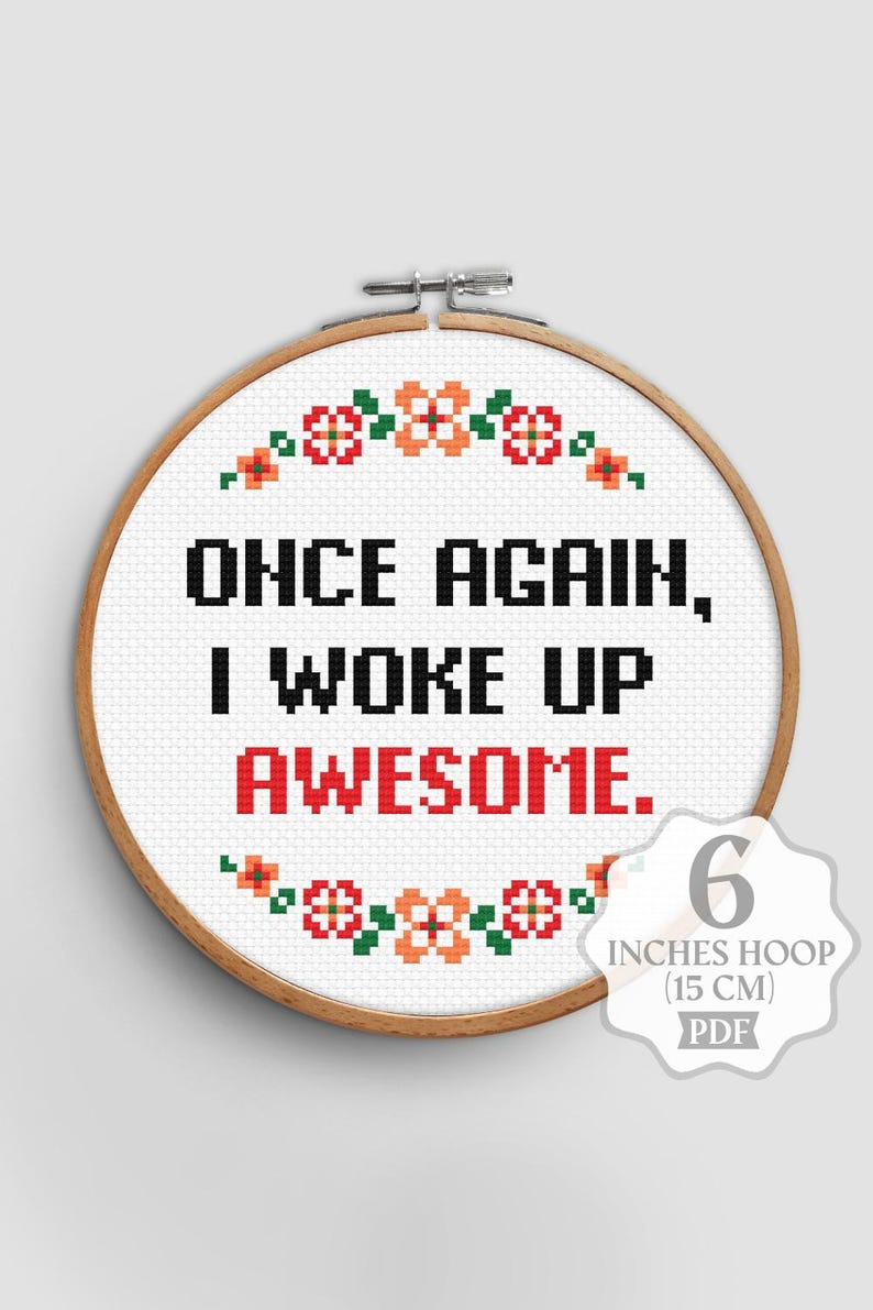 Op de afbeelding: Een borduurring met de tekst "ONCE AGAIN, I WOKE UP AWESOME." in zwart en rood. Het ontwerp bevat bloempatronen in oranje, rood en groen. De ring heeft een diameter van 15 cm.