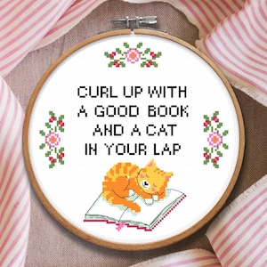 Op de afbeelding: Een ronde houten borduurring met witte stof en een kruissteekontwerp. De tekst luidt CURL UP WITH A GOOD BOOK AND A CAT IN YOUR LAP. Een oranje cyperse kat slaapt op een open boek, omlijst door kleine bloemmotieven.