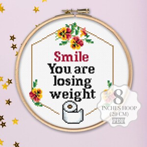 K&ouml;nnte beinhalten: Kreuzstichmuster mit einer Toilettenpapierrolle mit dem Text "Smile You are losing weight" in einem wei&szlig;en Rahmen mit roten und gelben Blumen. Das Muster ist f&uuml;r einen 20 cm gro&szlig;en Reifen.