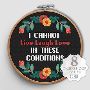 Op de afbeelding: Zwarte kruissteek in een borduurring met de tekst "I CANNOT Live Laugh Love IN THESE CONDITIONS". Het ontwerp bevat bloemmotieven in rood, geel en groen. De ring is 20 cm in diameter.