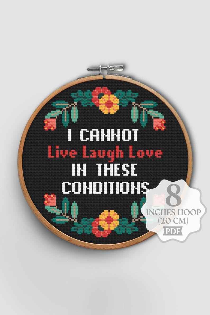 Op de afbeelding: Zwarte kruissteek in een borduurring met de tekst "I CANNOT Live Laugh Love IN THESE CONDITIONS". Het ontwerp bevat bloemmotieven in rood, geel en groen. De ring is 20 cm in diameter.