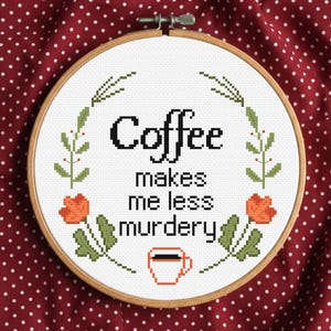 May include: A circular wooden embroidery hoop containing white fabric with a cross-stitch design. The text reads "Coffee makes me less murdery" in black, surrounded by green leafy stems, orange flowers, and a small orange coffee cup icon.