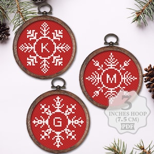 Puede incluir: Tres adornos de punto de cruz rojo y blanco con diseños de copos de nieve y una letra en el centro. Los adornos están en aros de 7,5 cm. Las letras son K, M y G.