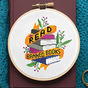 Op de afbeelding: Kruissteekborduurwerk in een houten ring met de woorden "READ BANNED BOOKS" bovenop een stapel boeken. Het ontwerp bevat oranje, paarse, groene en blauwe elementen, tegen een witte achtergrond. De borduursel is geplaatst op een bordeauxrood boek.