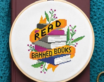 May include: Cross-stitch embroidery in a wooden hoop with the words "READ BANNED BOOKS" above a stack of books. The design includes orange, purple, green, and blue elements, set against a white background. The embroidery is placed on a maroon book.