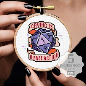 May include: Cross-stitch embroidery in a wooden hoop. The design features a purple and white die with the text "Crying is a Free Action" on a banner. The hoop is 12 cm in diameter.