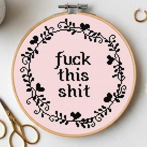 May include: A circular wooden embroidery hoop containing light pink fabric with a black cross-stitch design. The design features a floral wreath border surrounding the black text "fuck this shit" in a simple, block-style font centered on the fabric.