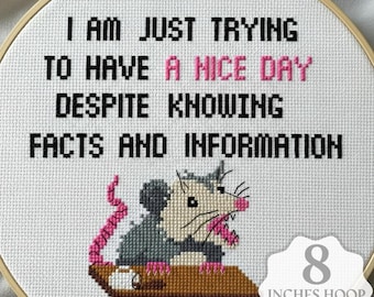 Könnte beinhalten: Ein Kreuzstichmuster in einem 20 cm großen Holzrahmen mit einem grauen und weißen Opossum mit rosa Nase und Schwanz. Der Text lautet: I AM JUST TRYING TO HAVE A NICE DAY DESPITE KNOWING FACTS AND INFORMATION auf einem weißen Stoffgrund.