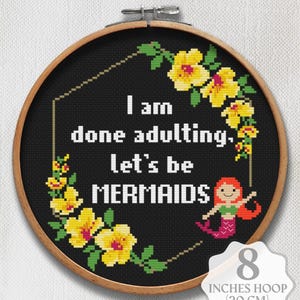 May include: A cross-stitch pattern in an 8-inch wooden hoop featuring the white text I am done adulting, let's be MERMAIDS on a black background, framed by yellow hibiscus flowers and a small mermaid with red hair and a pink tail.