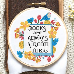 Op de afbeelding: Een ronde houten borduurring met witte stof en een kruissteekontwerp. De tekst BOOKS are ALWAYS A GOOD IDEA staat in het zwart, omringd door een kleurrijke bloemenrand van gele, roze en blauwe bloemen en een blauwe vogel.
