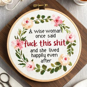 May include: A circular wooden embroidery hoop containing white fabric with a cross-stitch design. The text reads: A wise woman once said fuck this shit and she lived happily even after. The text is surrounded by a floral wreath of pink and peach roses.