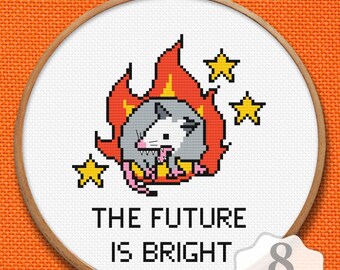 May include: A cross-stitch pattern in a 20 cm wooden hoop featuring a grey and white opossum surrounded by red and orange flames and three yellow stars. The text below the opossum reads THE FUTURE IS BRIGHT. The design is on white fabric.