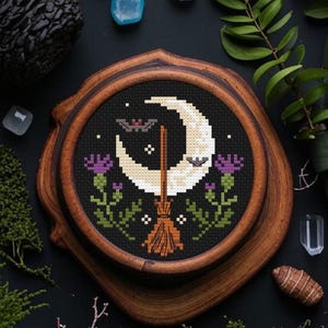 May include: A cross-stitch embroidery design featuring a crescent moon, two bats, a broom, and purple flowers on a black background. The design is framed in a wooden hoop.