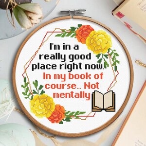 Op de afbeelding: Een kruissteekring met de tekst "I'm in a really good place right now. In my book of course... Not mentally." Het ontwerp bevat gele en oranje bloemen en groene bladeren. De ring is gemaakt van hout.