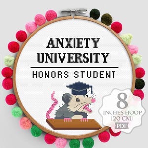 May include: A cross-stitch pattern in an 8-inch wooden hoop decorated with colorful pom-poms. The white fabric features the black text ANXIETY UNIVERSITY HONORS STUDENT and a pixelated opossum wearing a graduation cap on a brown desk.