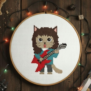 Puede incluir: Arte de punto de cruz en un aro de madera, que representa un gato de dibujos animados tocando una guitarra eléctrica roja. El gato tiene pelo castaño, una camisa azul y pantalones grises. El fondo incluye una vela, dados y luces de cadena.