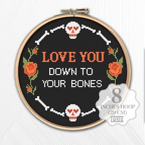 May include: A black cross-stitch embroidery hoop with the text "LOVE YOU DOWN TO YOUR BONES". The design includes white skulls, bones, and orange roses. The hoop is 20 cm in diameter.